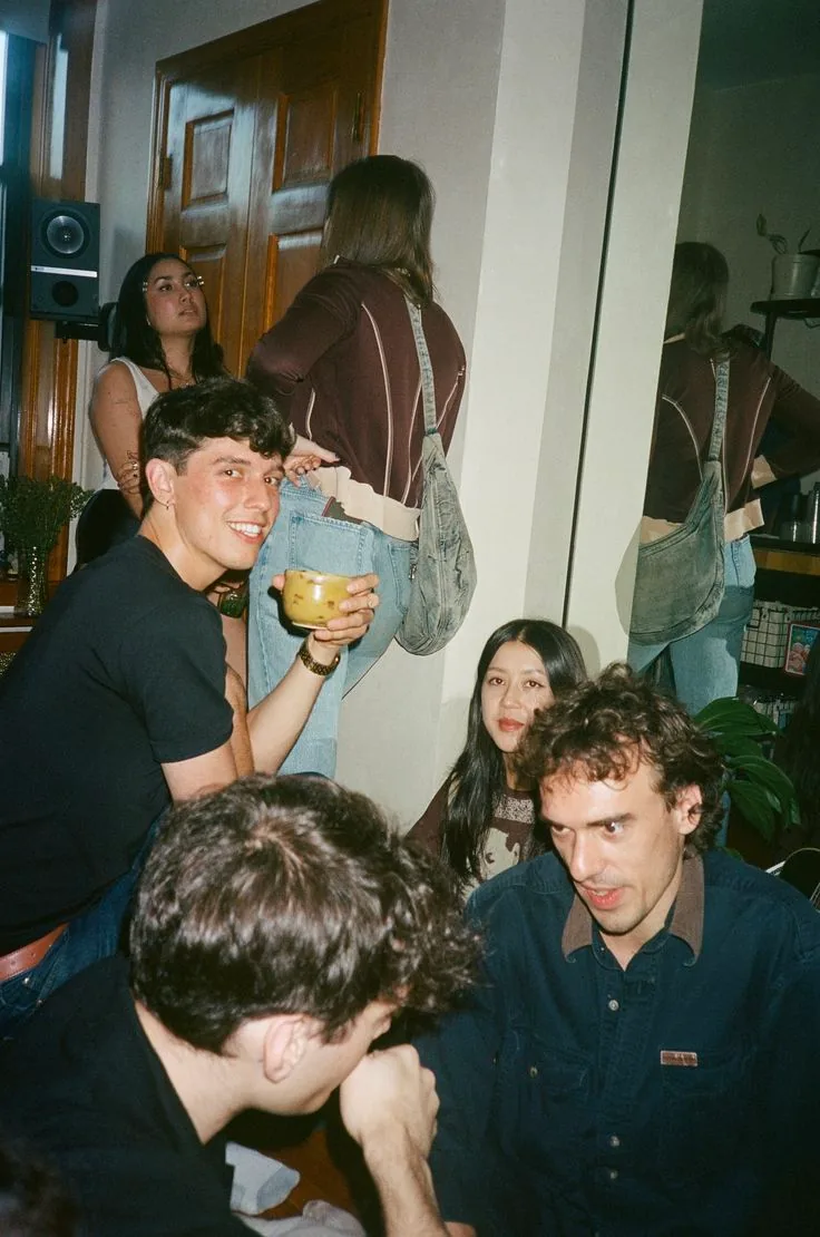 A crowded flat party in a hallway, a man turning to grin over his shoulder holding a mug.