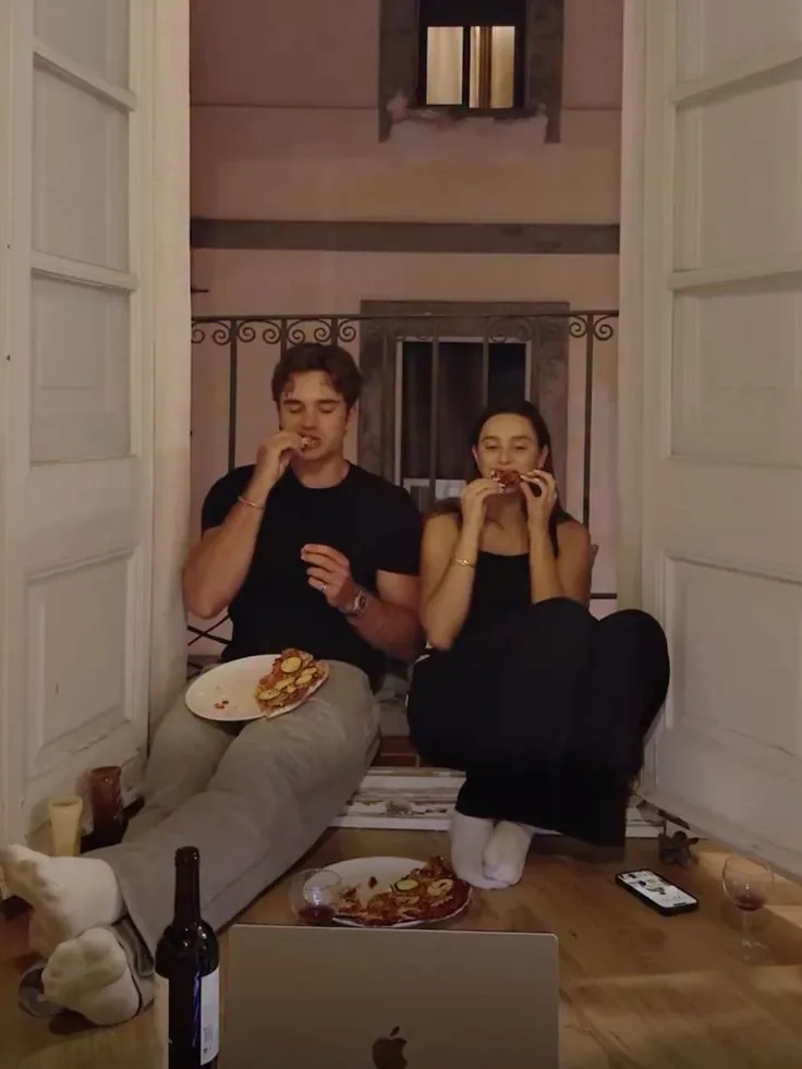 Two flatmates eating pizza on the floor by open balcony doors, a laptop open in front of them.