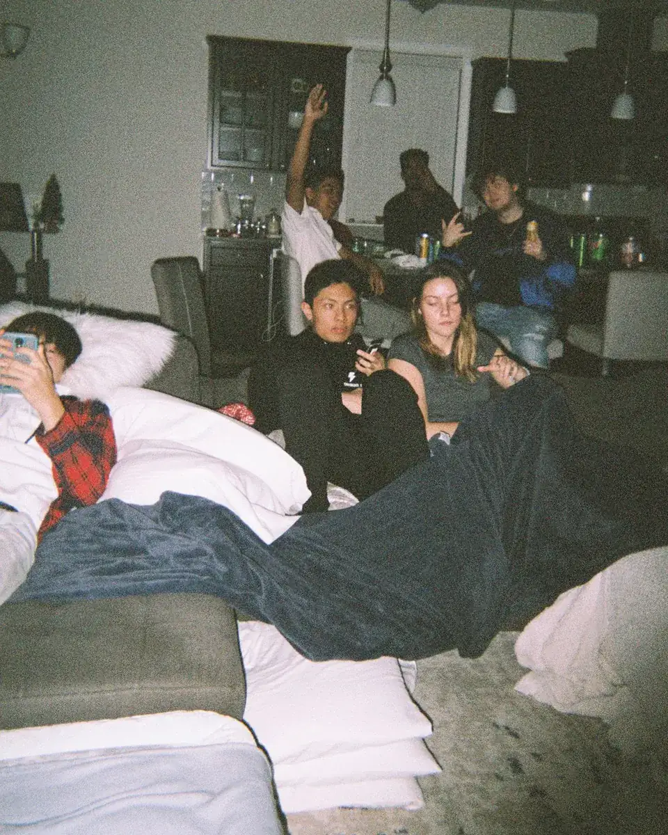 Six flatmates spread across a dark living room late at night with duvets pulled onto the floor.