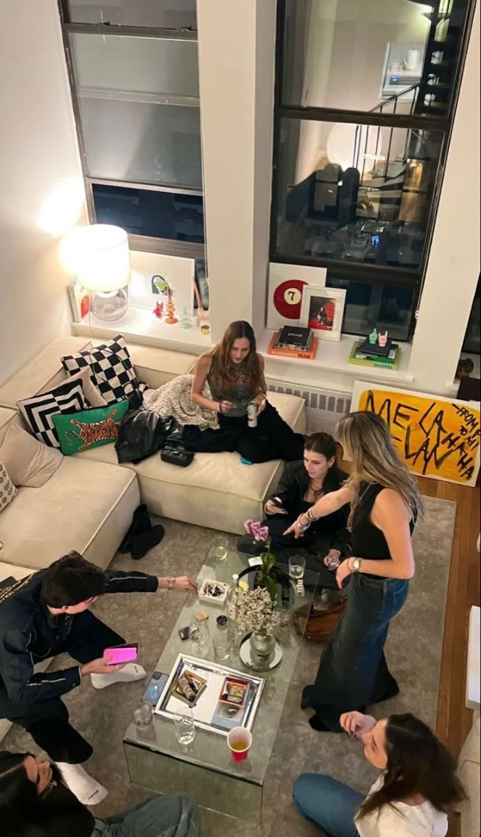 Seven friends around a low glass table in a tall-windowed flat, seen from above.
