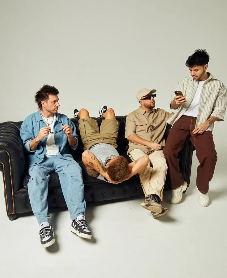 Four flatmates arranged along a black leather sofa against a plain backdrop, each doing something different.