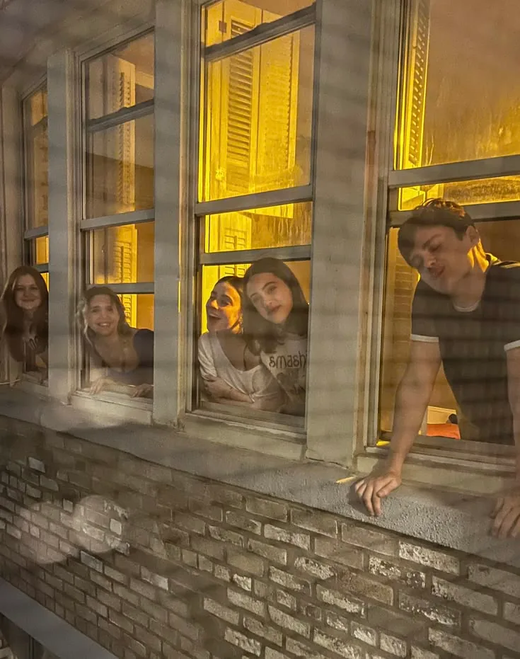 Four flatmates leaning out of lit windows at night, laughing down at the street.