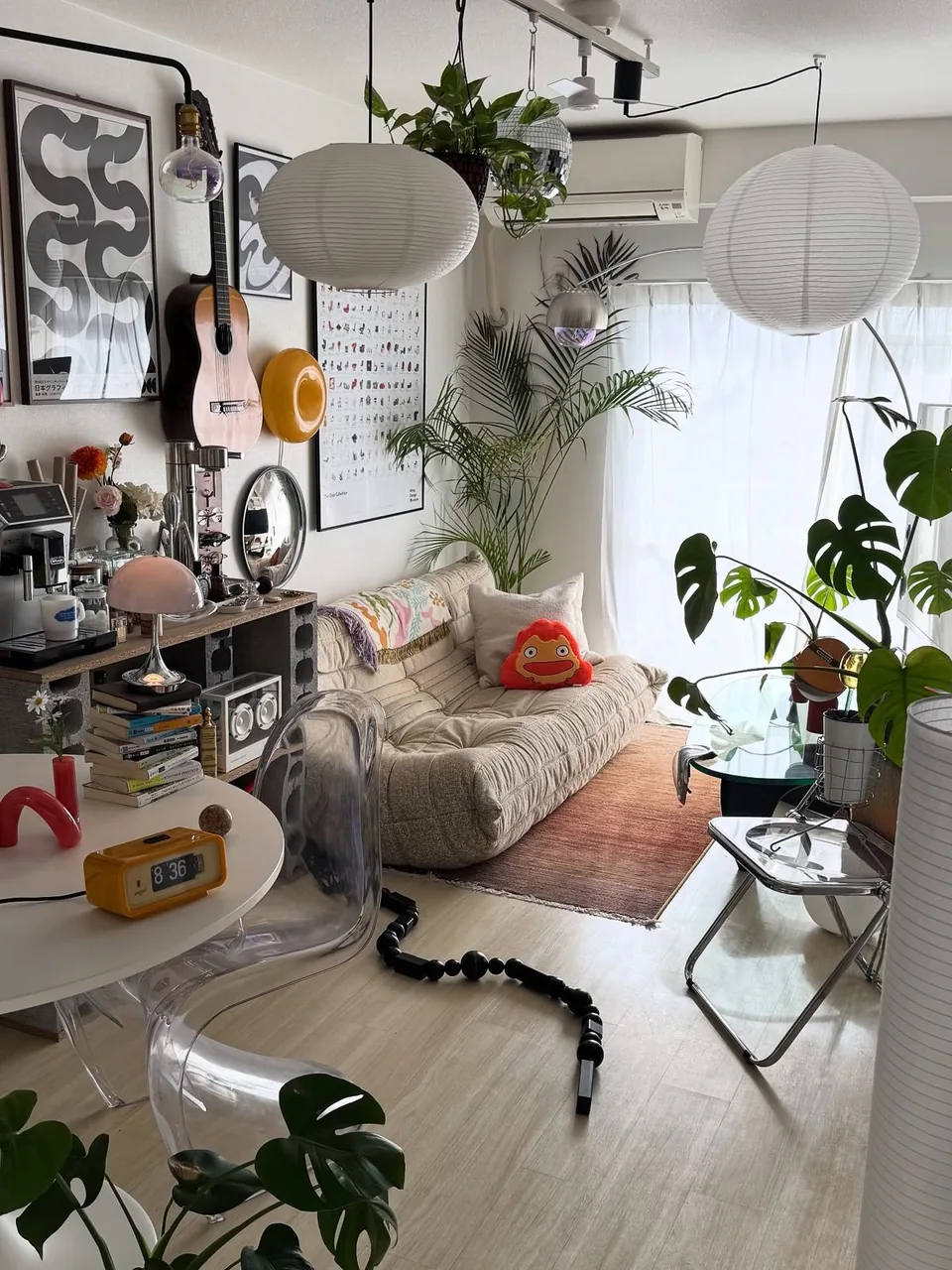 A small flat crowded with plants, paper lanterns and framed prints above a low sofa.